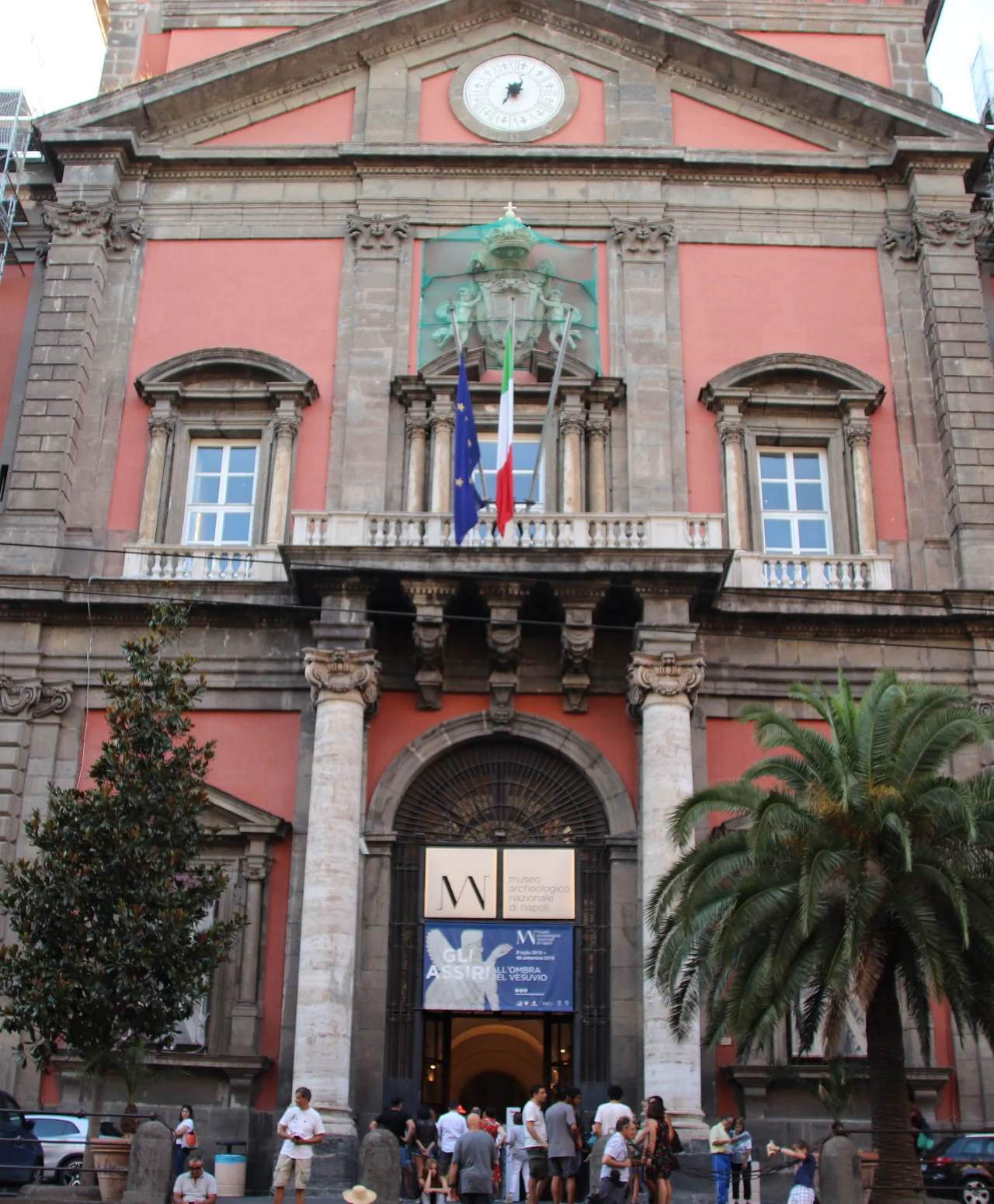 The red facade and arched main entrance of the National Archaeological Museum of Naples on Piazzale Museo, with museum banners and flags